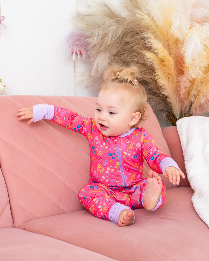 A baby girl wearing a pink floral long-sleeve pajama set sits on a dusty pink quilted sofa, with a white throw blanket and pampas grass in the background.