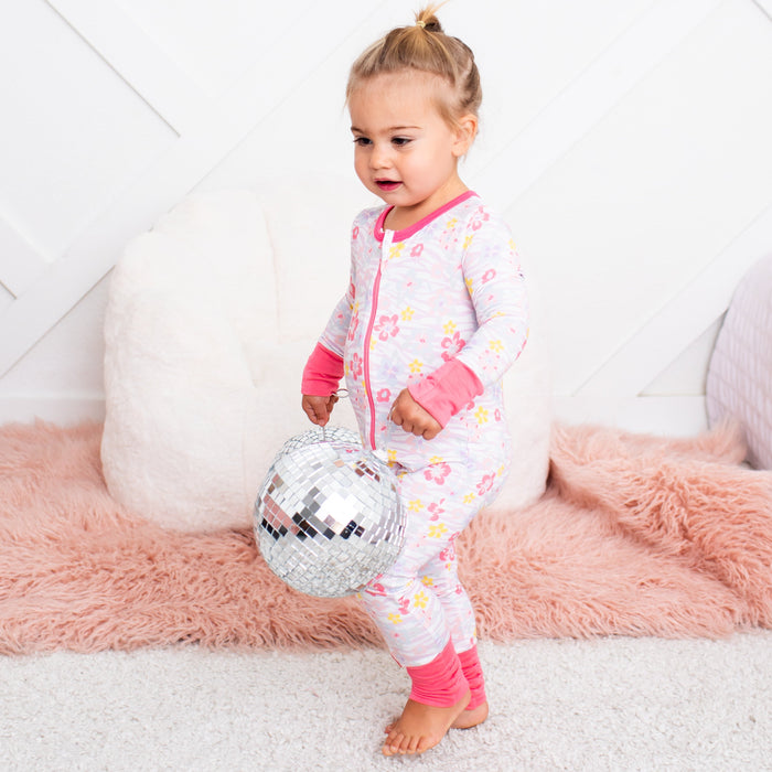 Child in a colorful onesie holding a disco ball on a white and pink rug.