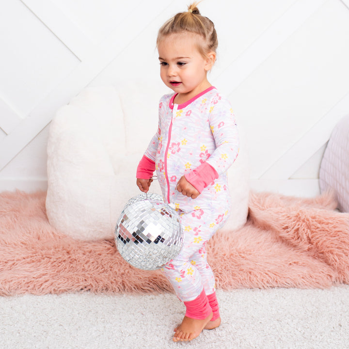 Child in a colorful onesie holding a disco ball on a white and pink rug.