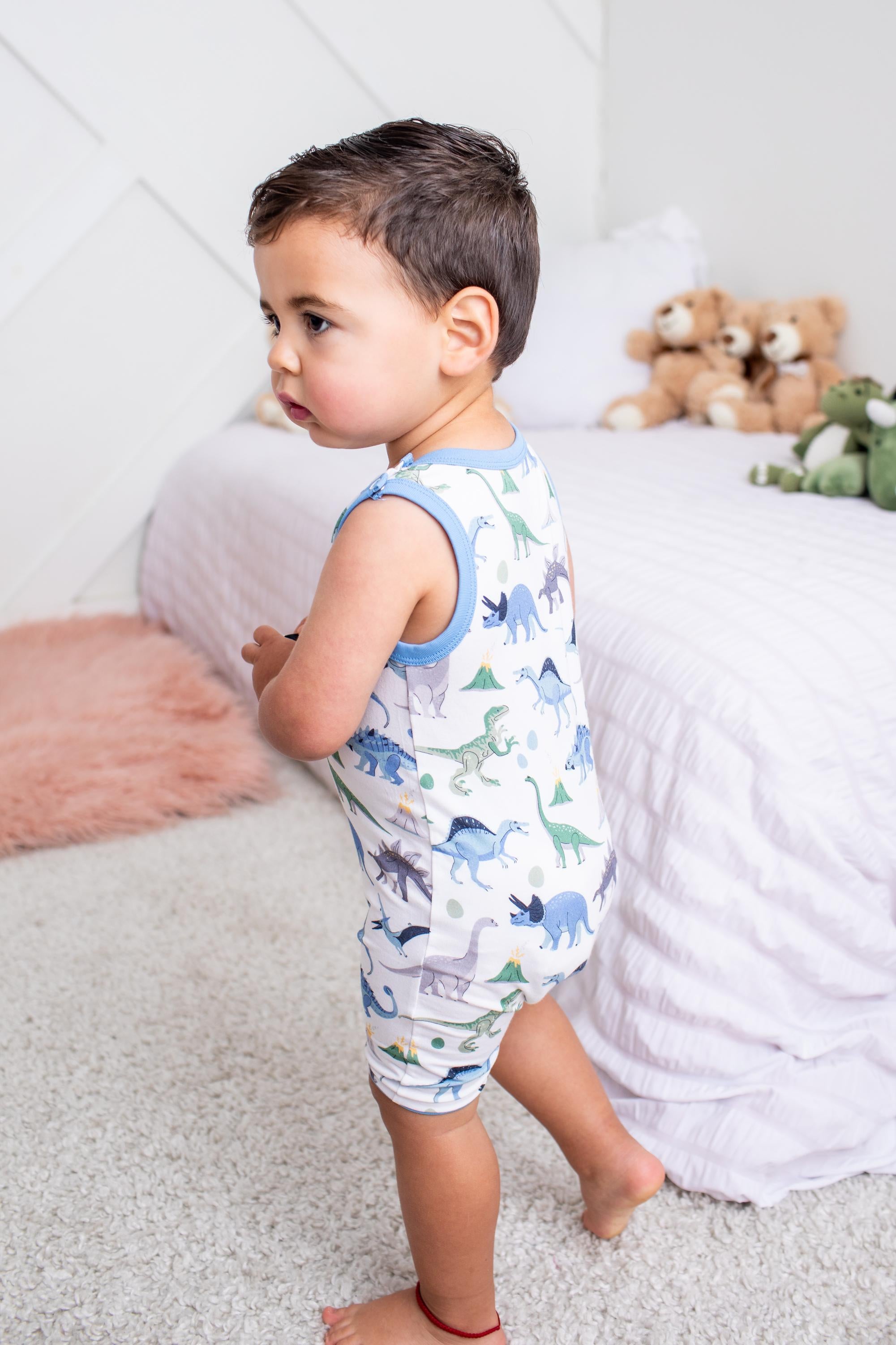 A toddler boy wearing a white sleeveless romper with a blue trim and a colorful dinosaur print, standing in a brightly lit bedroom.