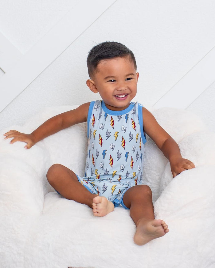 A toddler boy wearing a blue and white patterned sleeveless tank top and matching shorts sits smiling in a white plush armchair on a black and white checkered rug against a white paneled wall.