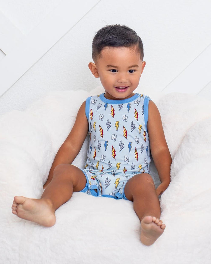 A young child sitting on a white plush bean bag chair, wearing a sleeveless light blue romper with a colorful fish pattern. The child is posed in front of a white paneled wall with a black and white checkered rug on the floor.