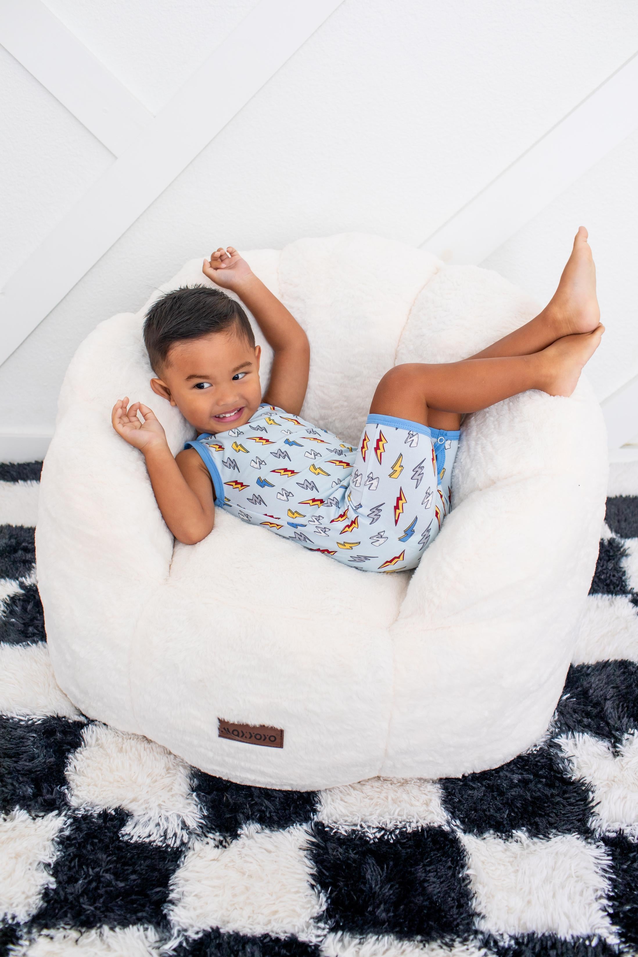 A young child wearing a light blue pajama set with a colorful fish pattern lies on their back inside a large, round, white plush bean bag chair. The chair is placed on a black and white checkered shag rug against a white paneled wall.