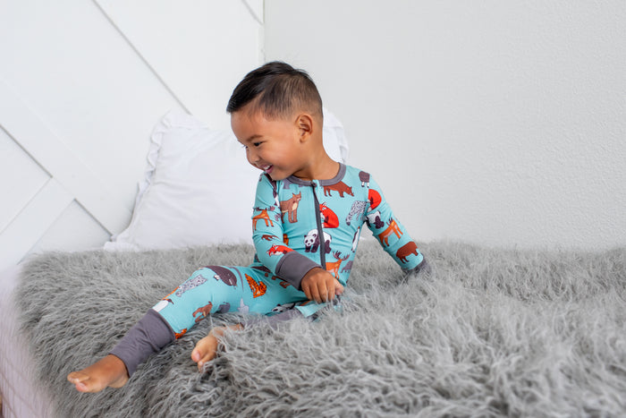 A young boy wearing a light blue long-sleeve pajama set with a colorful woodland animal print sits on a fluffy gray bedspread in a bright bedroom.