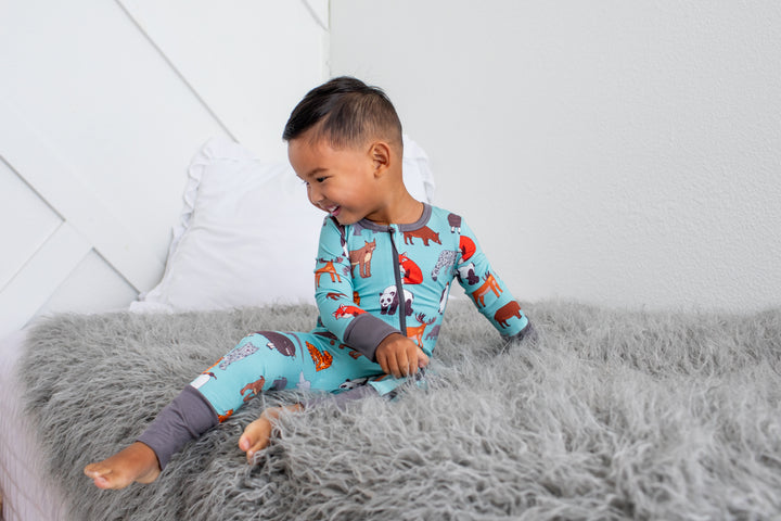 A young boy wearing a light blue long-sleeve pajama set with a colorful woodland animal print sits on a fluffy gray bedspread in a bright bedroom.