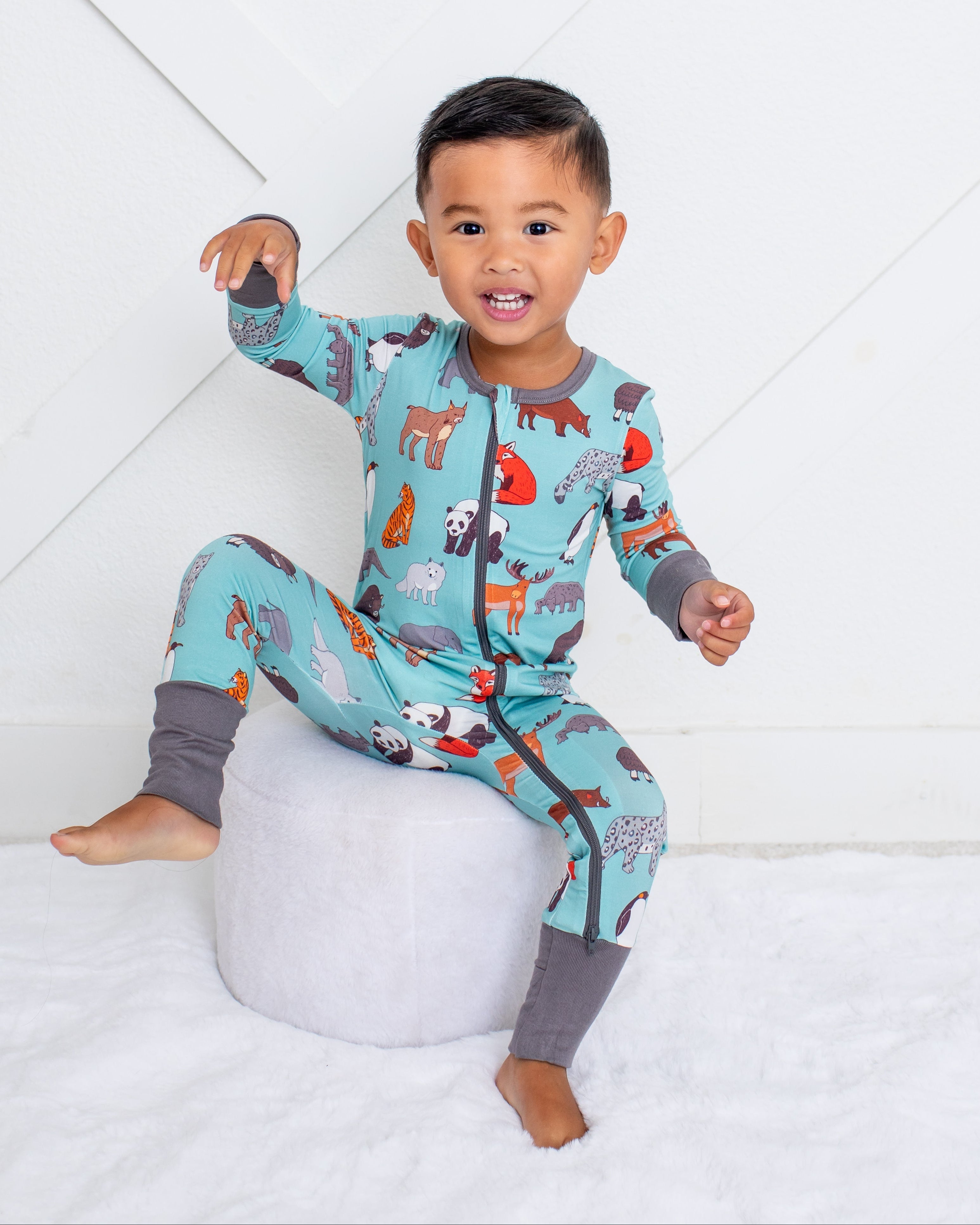A toddler boy wearing a light blue animal-print pajama set with gray leg warmers, sitting on a round white ottoman against a white paneled wall.