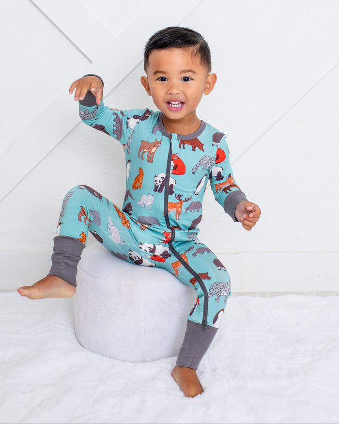 A toddler boy wearing a light blue animal-print pajama set with gray leg warmers, sitting on a round white ottoman against a white paneled wall.