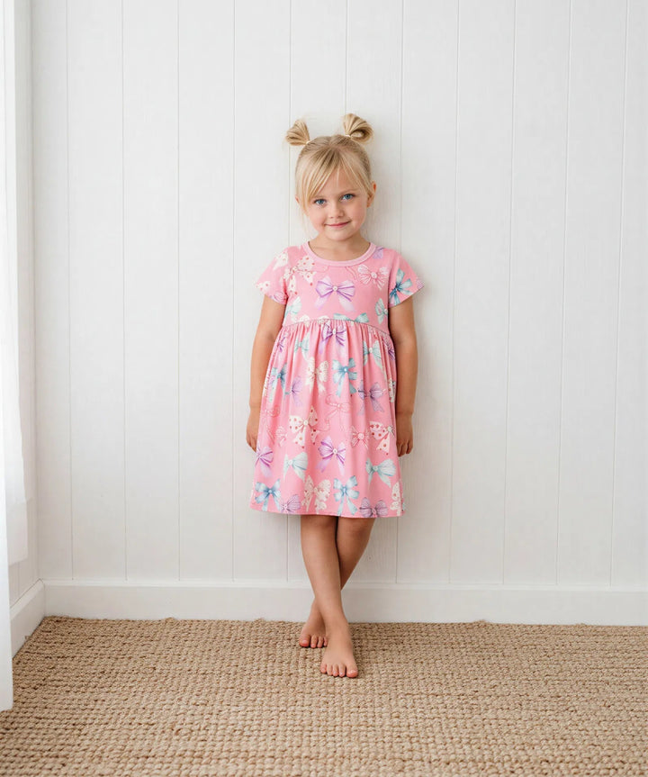Young girl in a pink floral dress standing against a white wall.