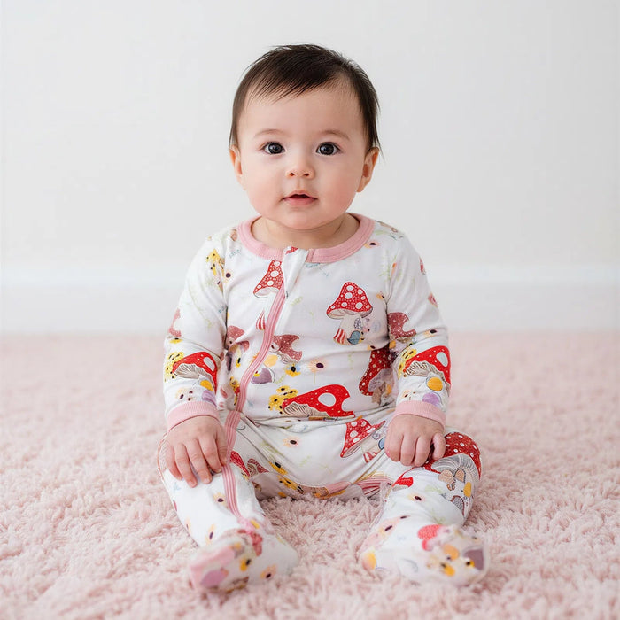 Baby sitting on a pink surface wearing a white onesie with colorful mushroom patterns.
