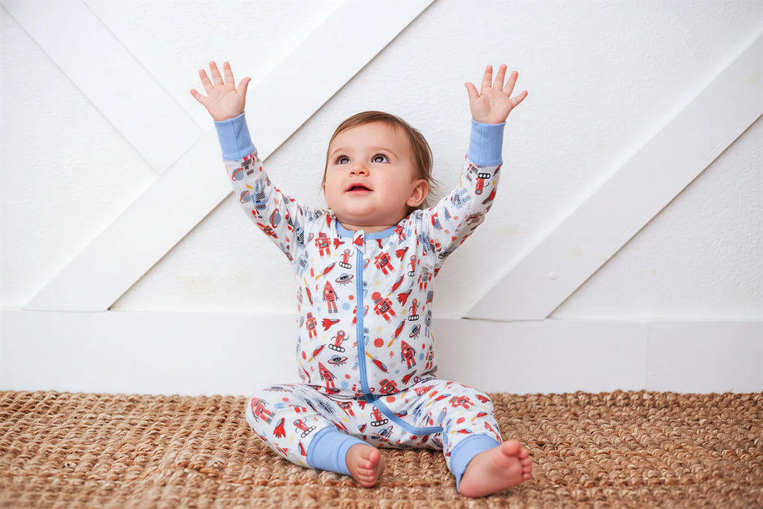 Baby wearing a colorful onesie with raised arms on a wooden floor.